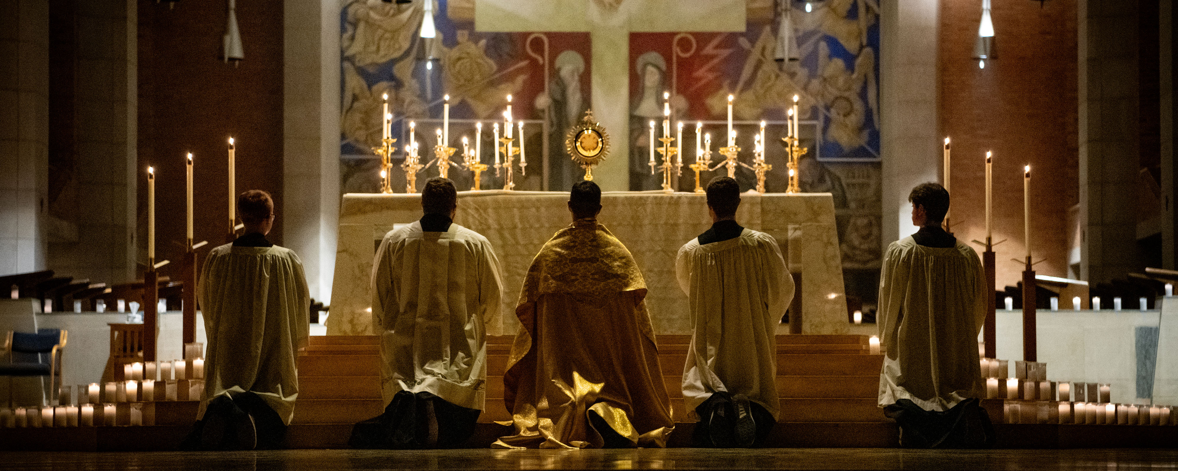 Priest and 4 altar servers kneeling in adoration with votive candles